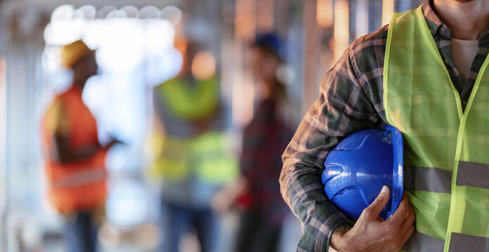 Man holding blue helmet close up. Construction man worker with office and people in background. Close up of a construction worker's hand holding working helmet.