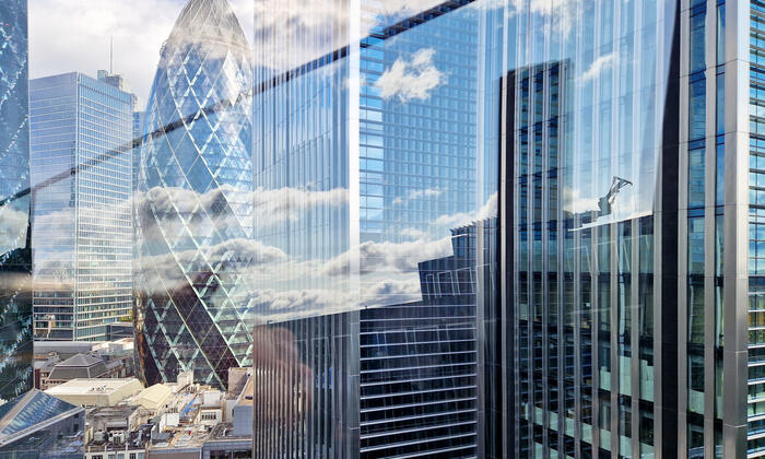 View of the Gherkin and surrounding modern glass skyscrapers in the City of London, with blue sky and clouds reflected in the mirrored façades and overlapping in the window glass, creating an abstract cityscape on a bright, partly cloudy day in the financial district.