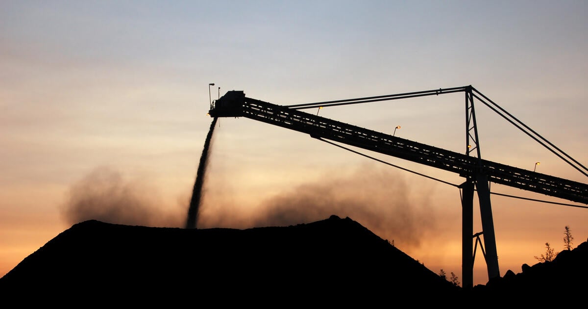 Industrial conveyor belt pouring dark material onto a large pile at sunset