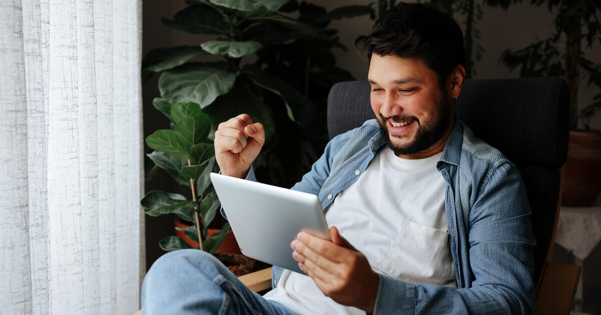 Male is sitting on sofa at home. He is in casuals. Shot of a happy young man using his tablet while relaxing on the couch at home