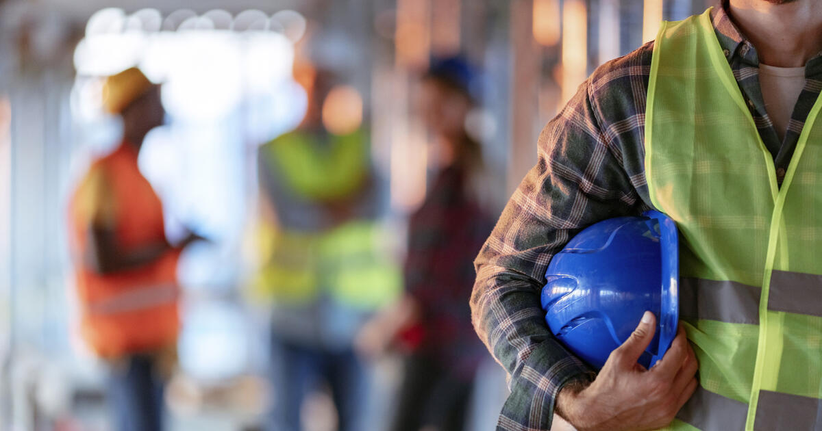 Man holding blue helmet close up. Construction man worker with office and people in background. Close up of a construction worker's hand holding working helmet.